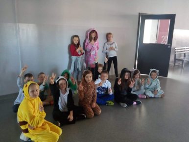 Zdjęcie przedstawia grupę uczniów siedzących wspólnie w holu szkoły, ubranych w kolorowe, pluszowe piżamy, kapcie i szlafroki.